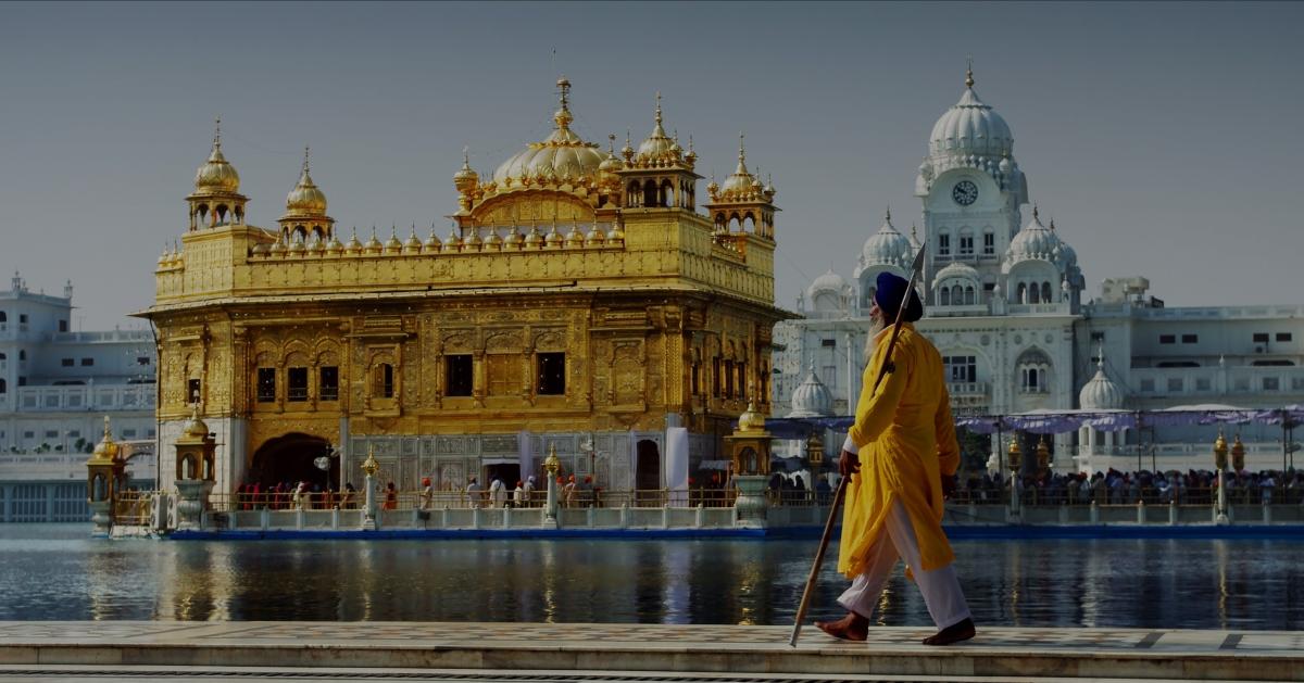 En sikhmand i gul kappe og med spyd går langs vandet, i baggrunden den Gyldne Tempel i Amritsar, Ind